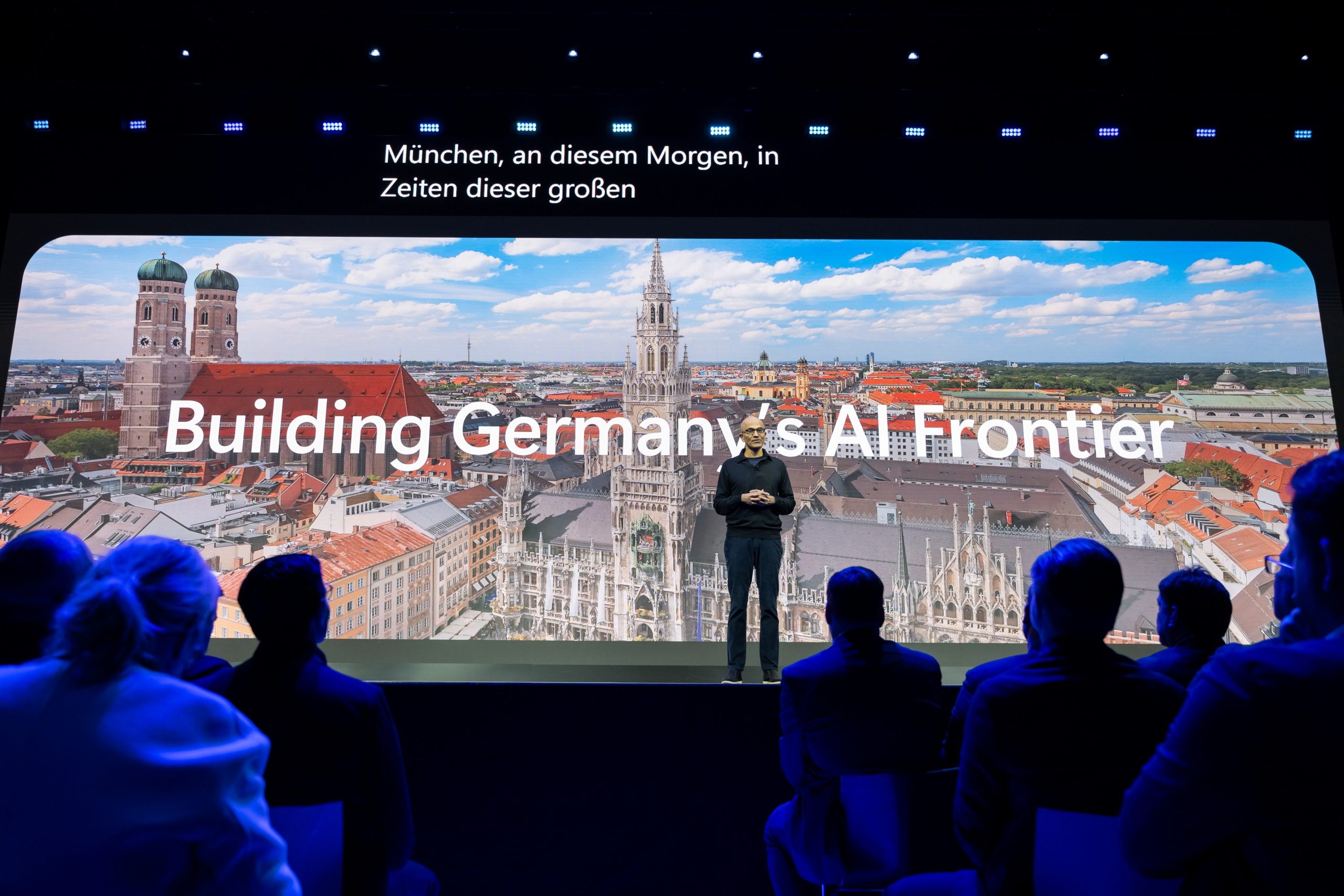 Redner auf der Bühne vor Münchner Skyline und dem Schriftzug „Building Germany’s AI Frontier“ mit Publikum im Vordergrund.