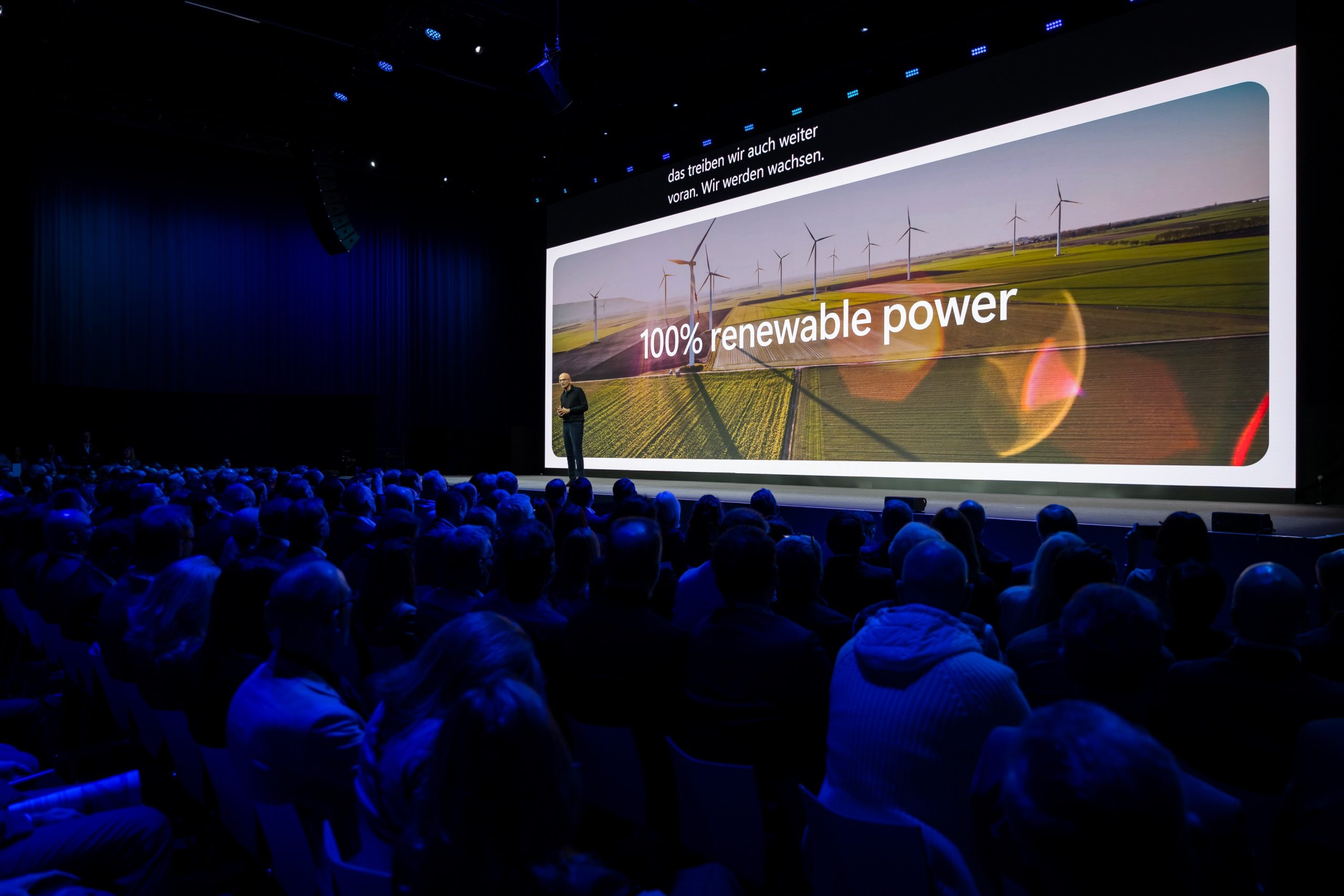 Saal mit großem Publikum; auf der Bühne präsentiert eine Person vor einer großen Leinwand mit Windrädern und der Überschrift „100% renewable power“.