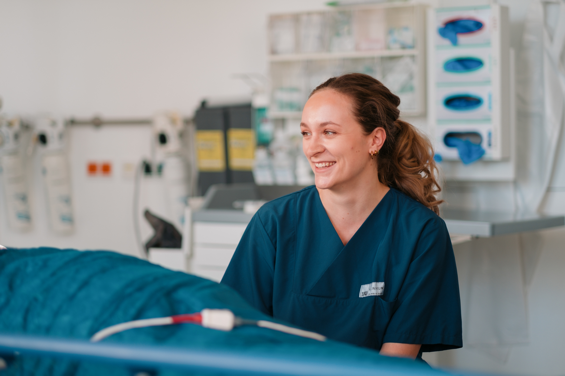 Porträt einer lächelnden Frau in blauer OP-Kleidung, die neben einer Krankentrage mit dem Umriss eines Patienten sitzt.