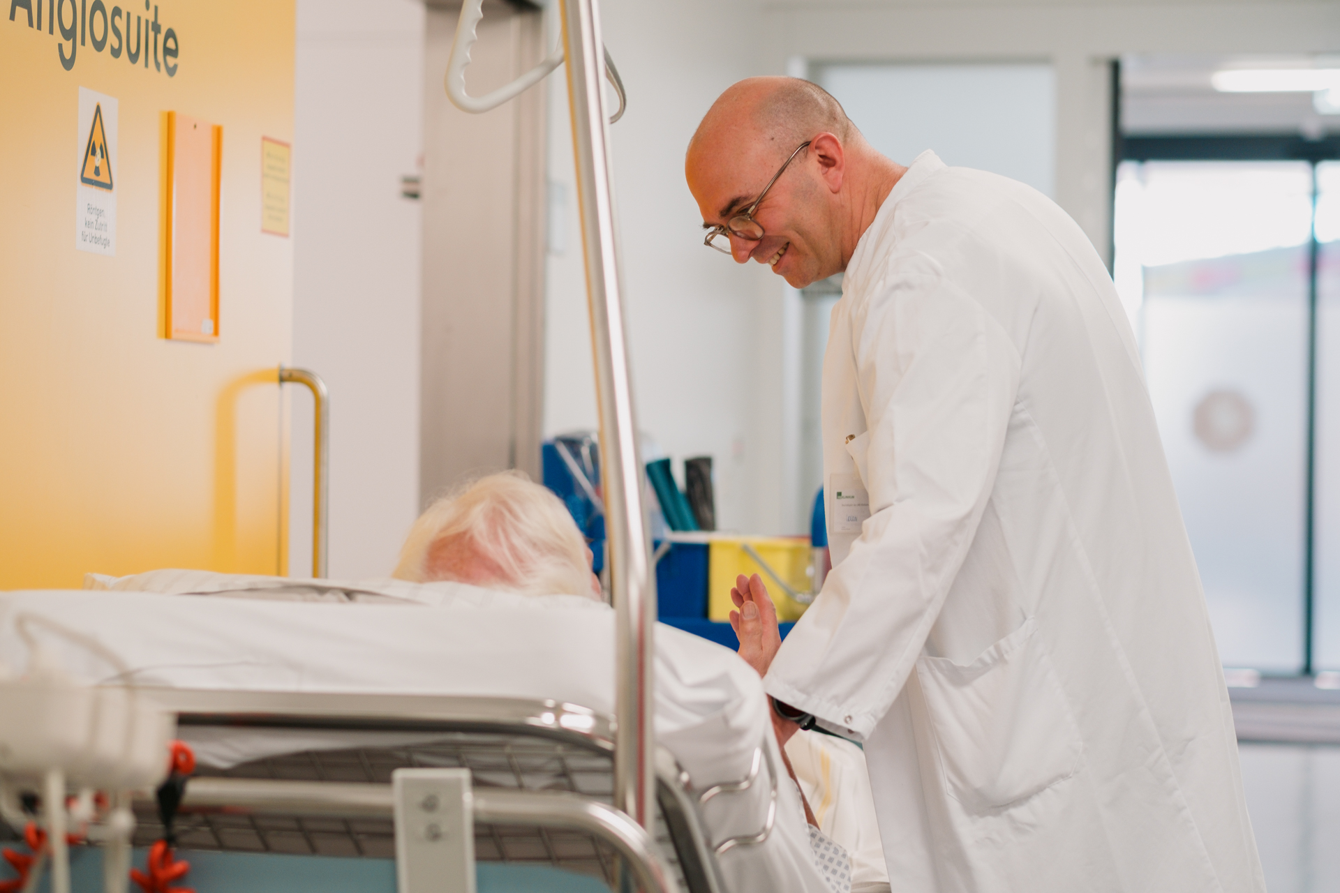 Mann in weißem Laborkittel mit Brille, der sich über einen Patienten (nur die Haare sind sichtbar) auf einer Trage beugt.
