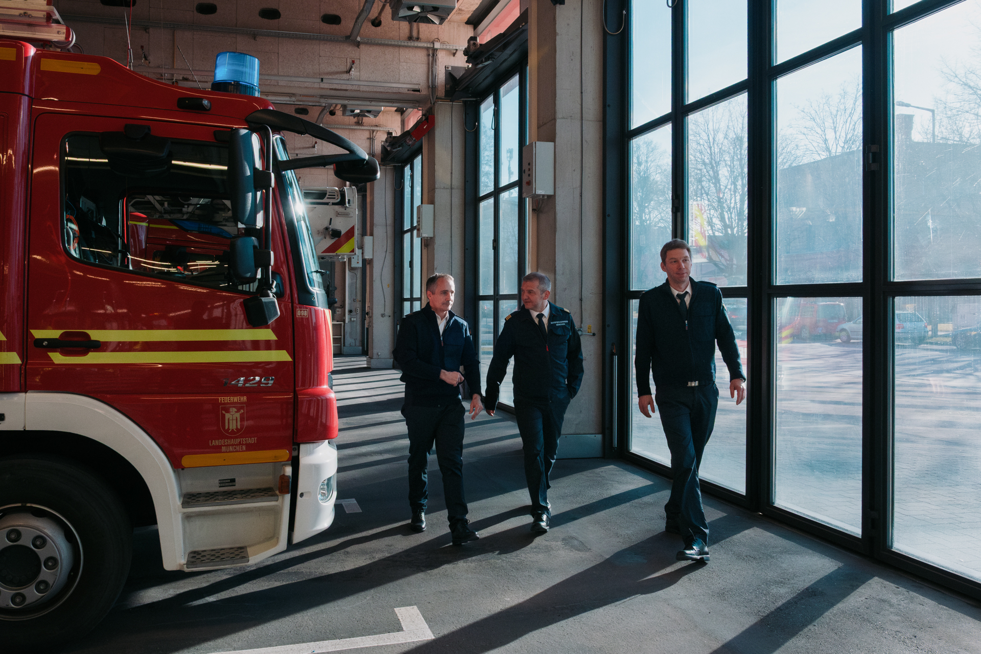 Drei Männer in Uniform gehen in der sonnendurchfluteten Halle einer großen Garage mit Feuerwehrautos auf die Kamera zu.