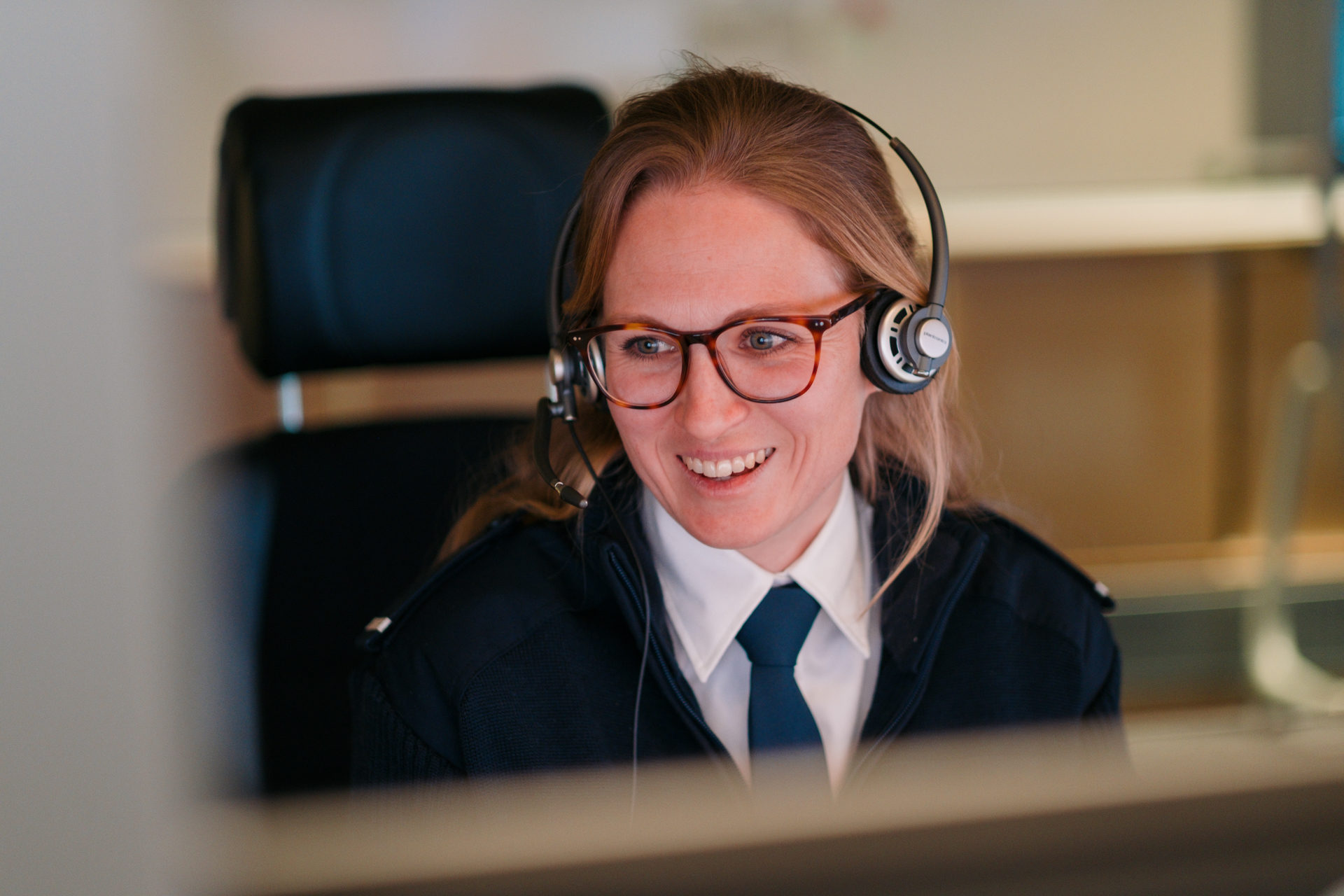 Lächelnde Frau mit Brille und Telefon-Headset an einem Computerarbeitsplatz.