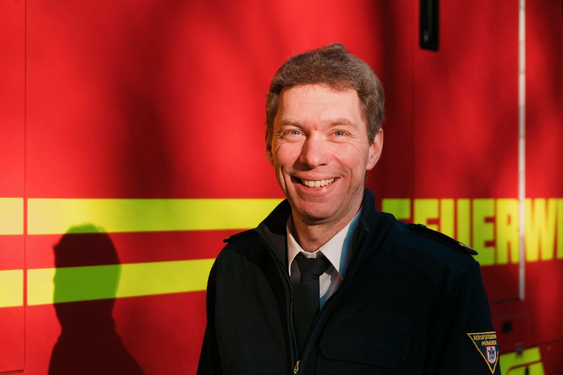 Porträt eines lächelnden Mannes in Feuerwehruniform vor einem Teil eines roten Feuerwehrautos.