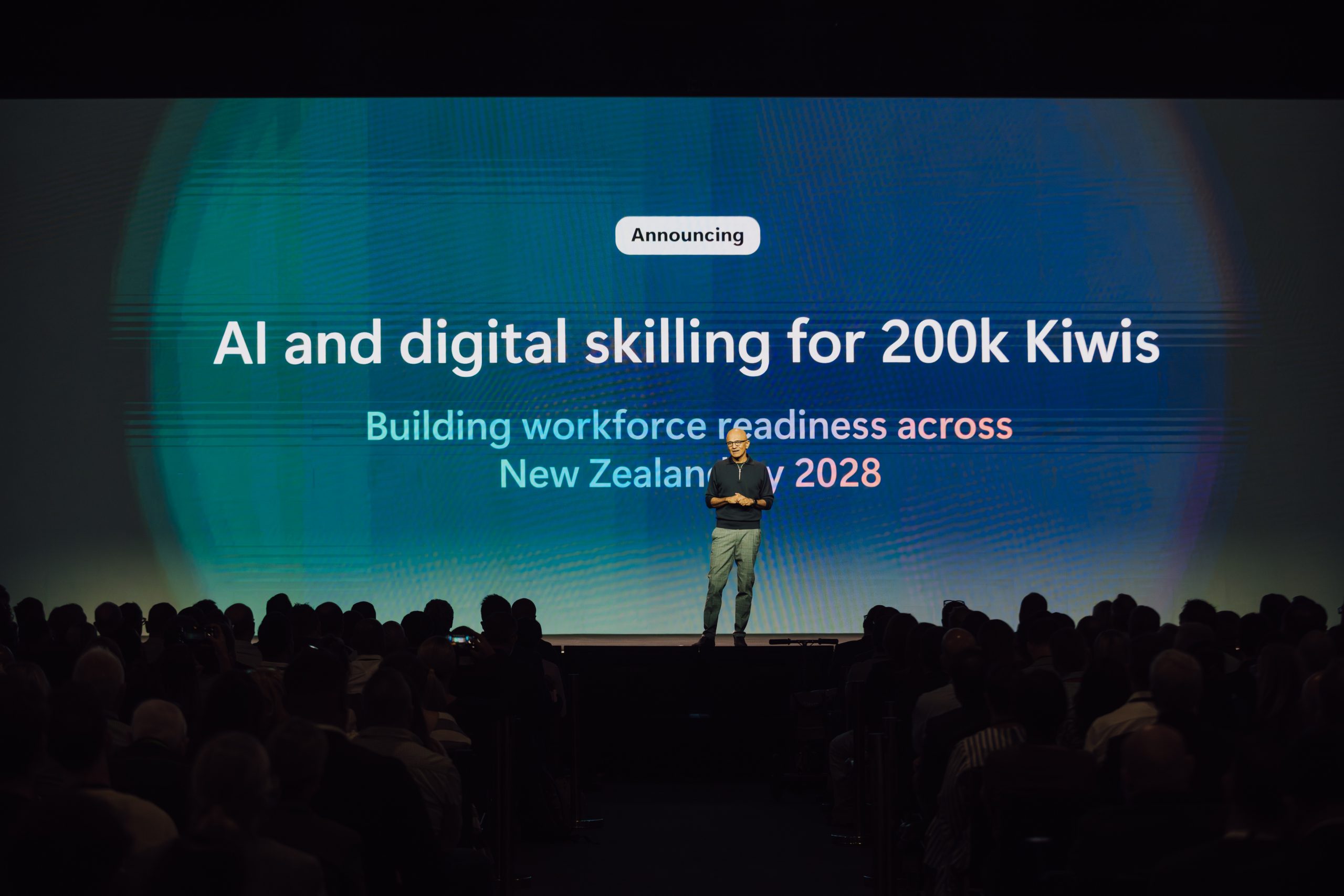 a man in a black sweatshirt on stage making a presentation, backdrop reads 'AI and digital skilling for 200K Kiwis'